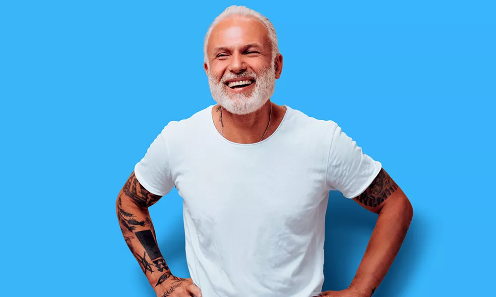 Man met wit shirt grijs haar en tatoeages, blauwe achtergrond